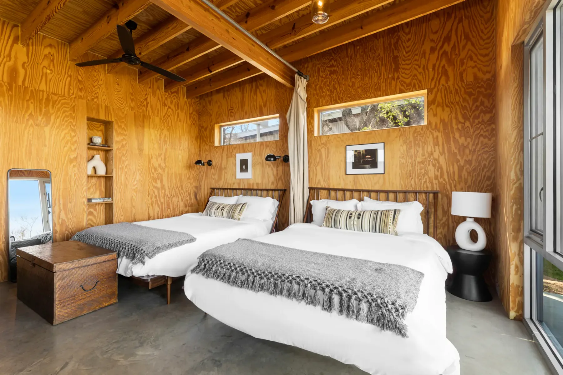 Spacious bedroom with two beds, wooden walls and ceiling, large window with scenic view, wicker trunk, and a wooden desk with chair.