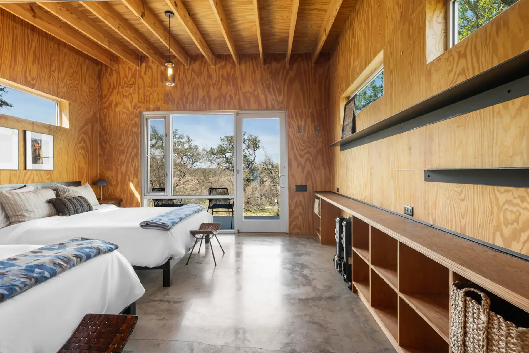 Modern bedroom with wooden walls, two beds, minimalist decor, large windows, and a glass door opening to a scenic view of trees outside.