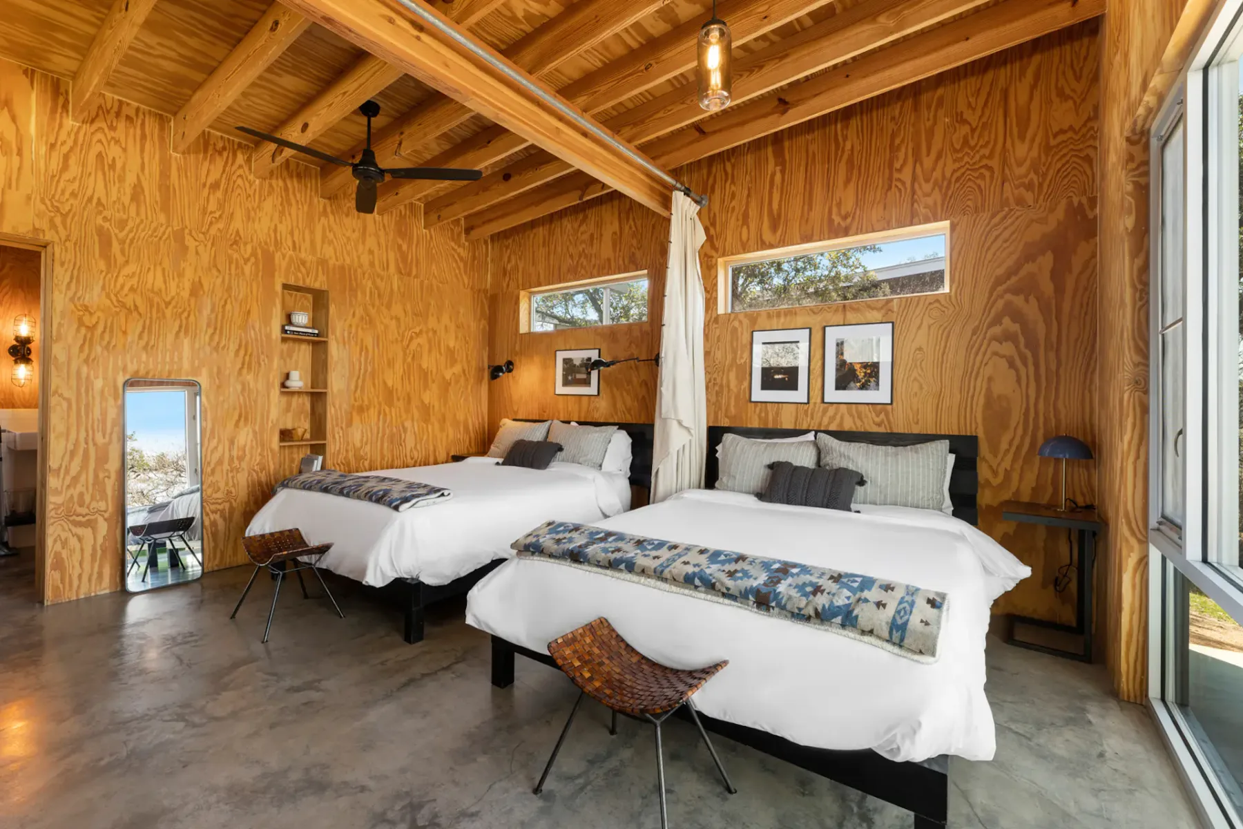 Cozy bedroom with two double beds, wooden walls, a ceiling fan, framed artwork, and a large window. Modern rustic decor.