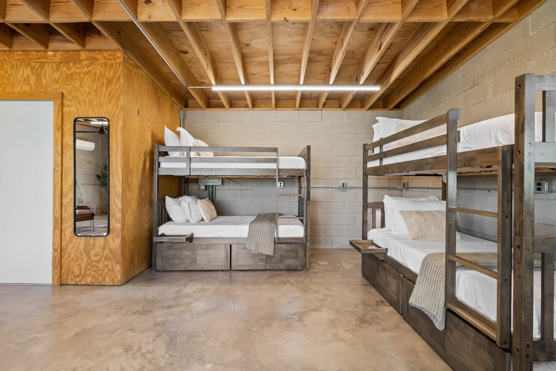 Room with two sets of wooden bunk beds, concrete floor, wooden ceiling, mirror on wall, and minimal decor.