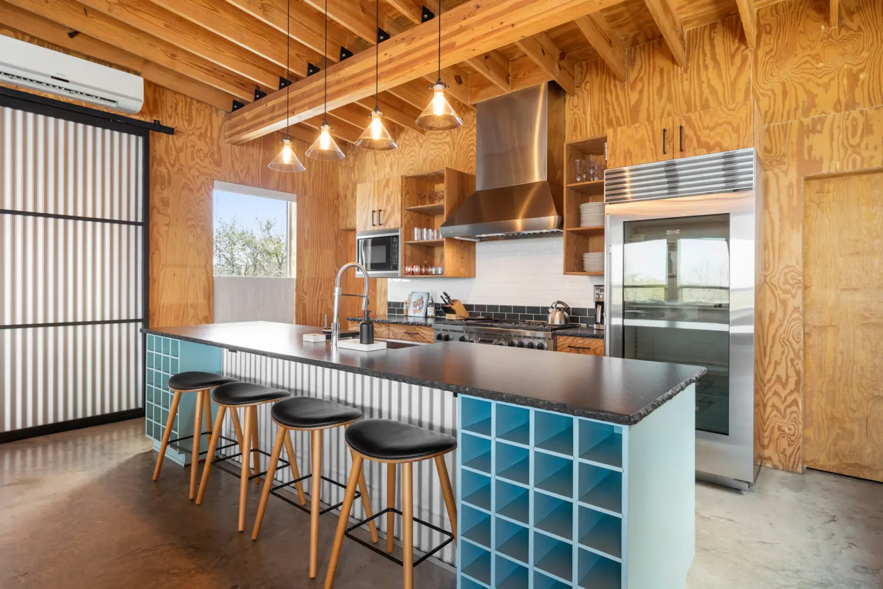 Modern kitchen with wooden walls, exposed beams, a large island with stools, wine rack, stainless steel appliances, and pendant lights.