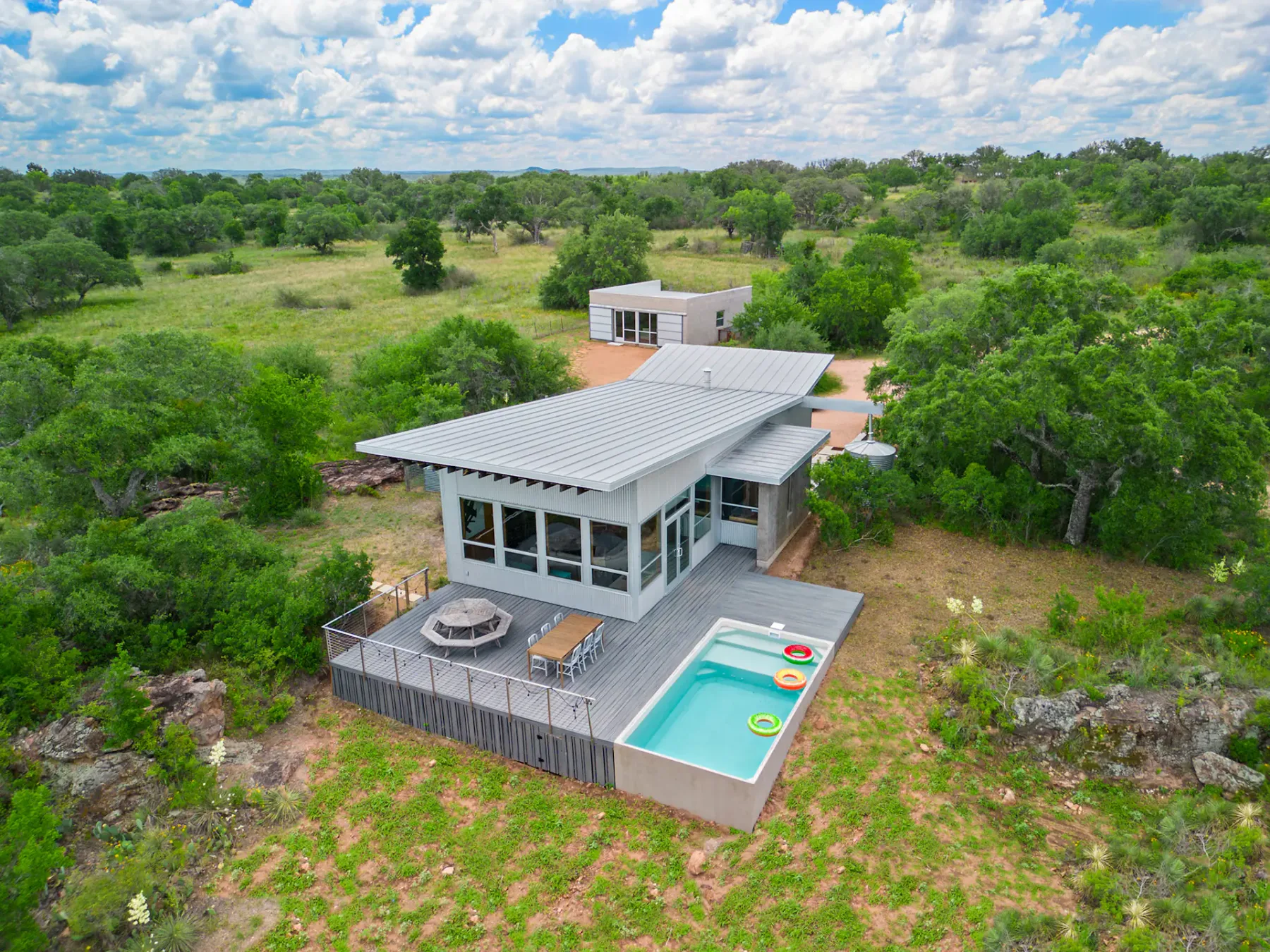 Modern house with large windows, a small pool, and outdoor seating, set in a lush green landscape under a partly cloudy sky.
