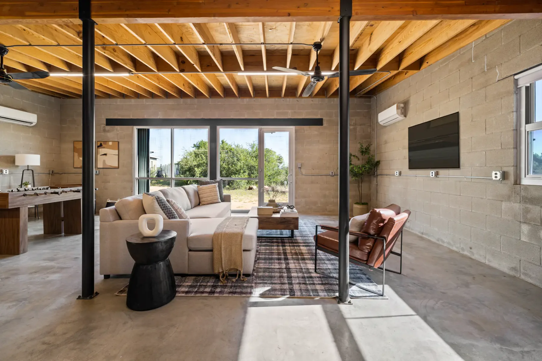 Spacious modern living room with wooden ceiling, sectional sofa, armchair, and large windows overlooking greenery. Foosball table in the corner.