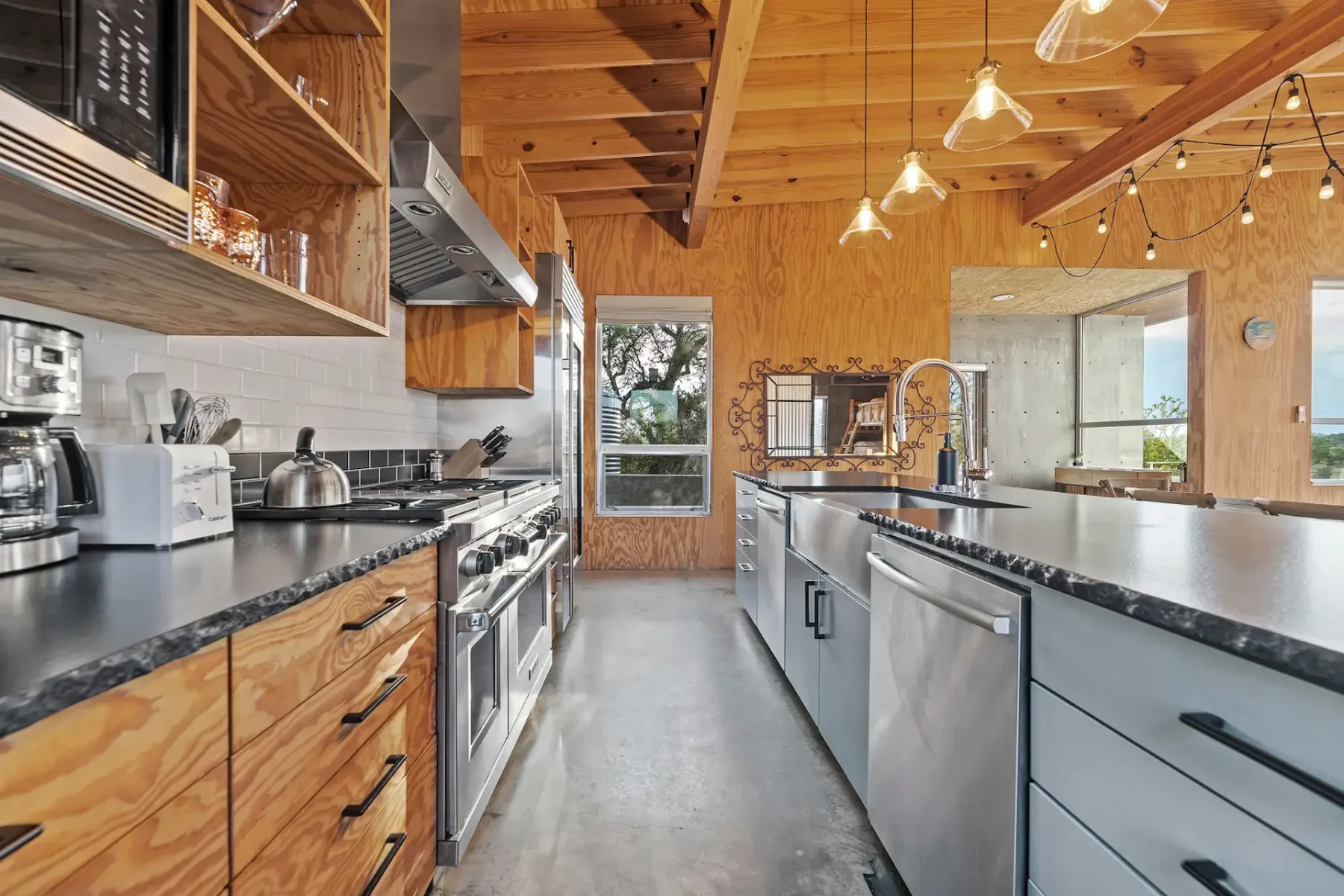 Modern kitchen with wooden cabinets, stainless steel appliances, black countertops, and pendant lights; large window with a scenic view.