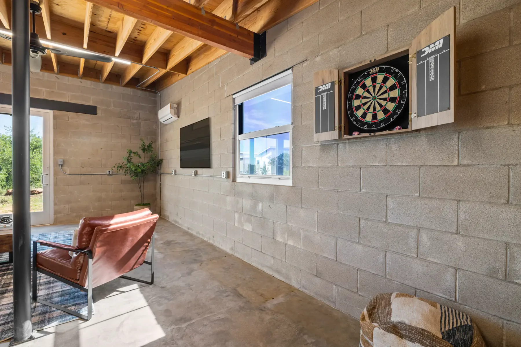 Room with concrete walls featuring a dartboard, leather chairs, a window, a chalkboard, and a basket. Large window provides natural light.