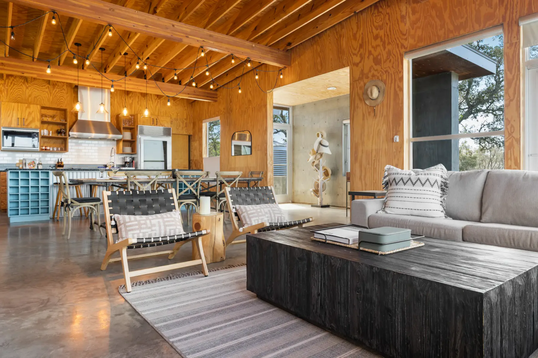 Open-plan living area with wooden walls, exposed beams, modern furniture, and large windows offering a view. Cozy seating and decorative elements.
