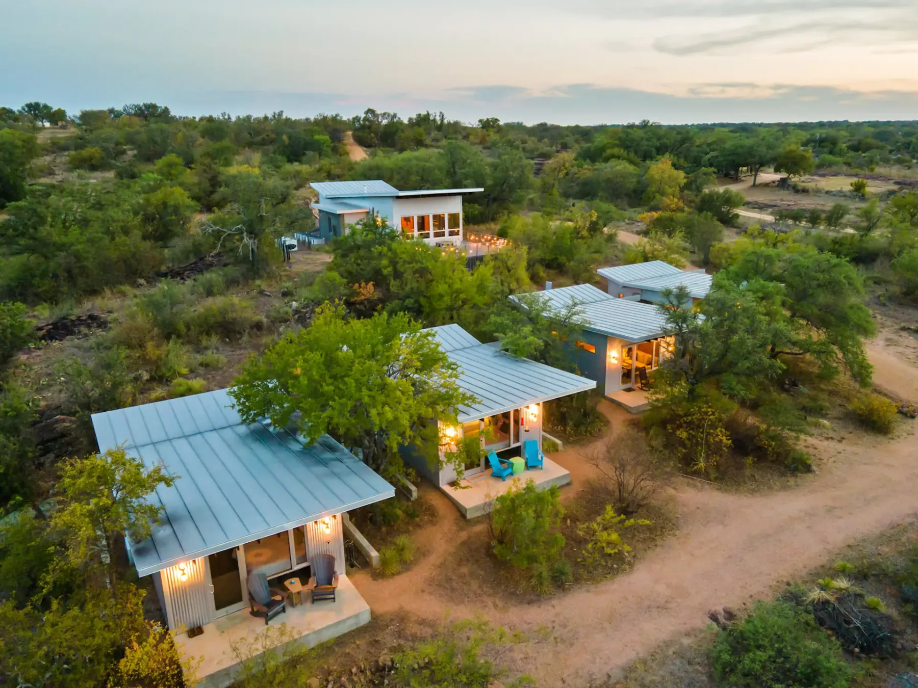 Aerial view of small cabins with metal roofs surrounded by trees and dirt paths during twilight in a rural setting.