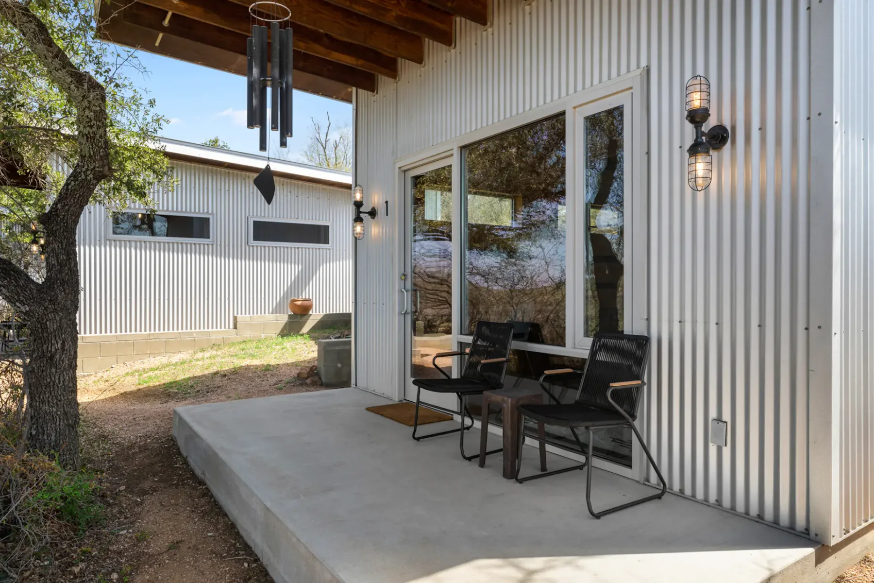 A small porch with two black chairs and a wind chime, attached to a modern building with corrugated metal siding and outdoor lights.