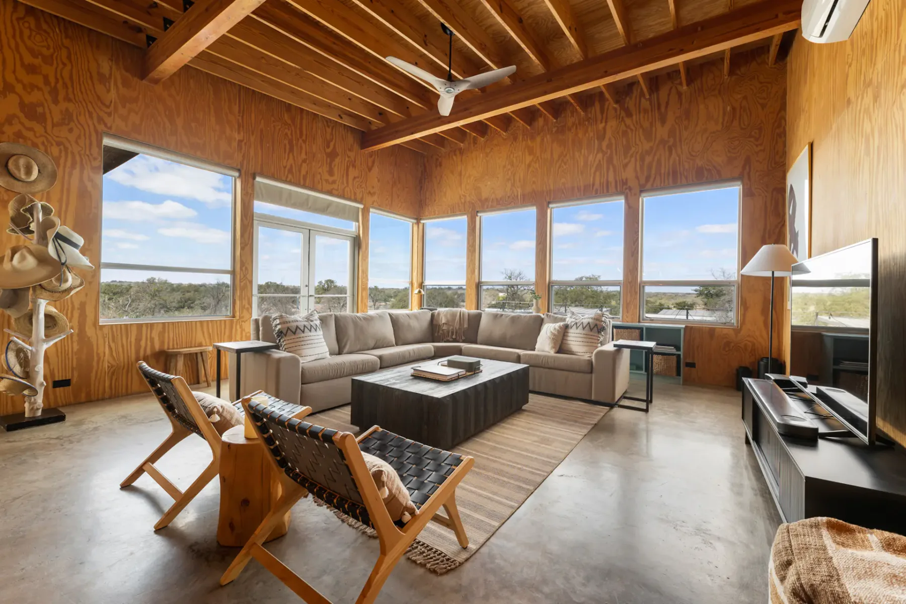 Modern living room with wooden walls, large windows, a sectional sofa, and woven chairs. Overlooks scenic outdoor view under a wooden beam ceiling.