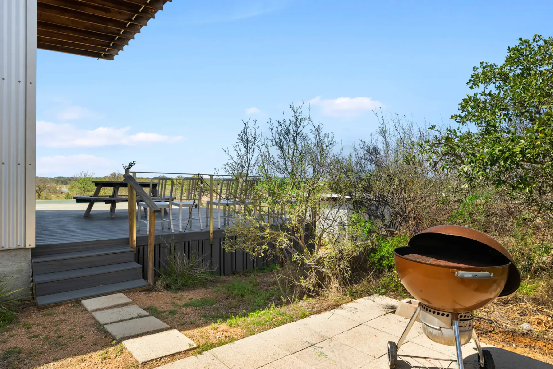 Patio with a wooden deck, picnic table, and chairs overlooking shrubs. Round charcoal grill on stone pavement in the foreground. Clear blue sky above.