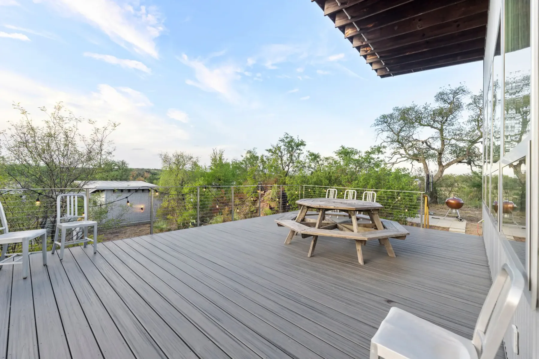 Spacious wooden deck with round picnic table and chairs, surrounded by trees and overlooking a clear sky.