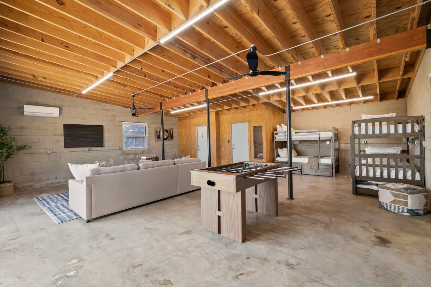 Spacious room with exposed wooden ceiling, sectional sofa, foosball table, bunk beds, and metal shelving on concrete floor.