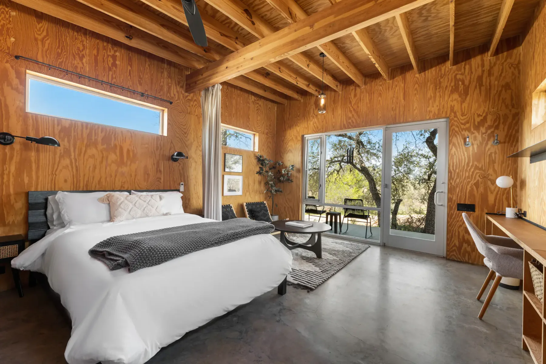 A cozy wooden bedroom with a large bed, desk, and seating area. Glass doors and windows offer natural light and a view of trees outside.