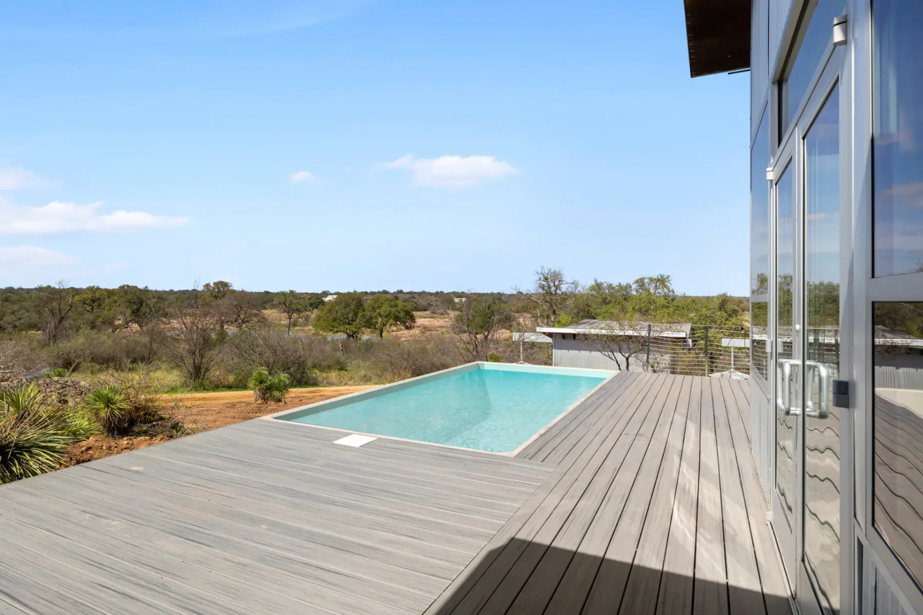Modern villa with large glass windows and a rectangular pool on a wooden deck, overlooking a vast, dry landscape under a clear blue sky.