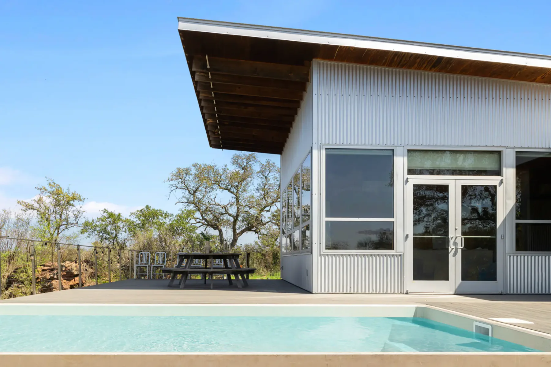 Modern house with large windows, metal siding, and sloped roof. Backyard features a pool, patio, and tree in the background under a clear sky.