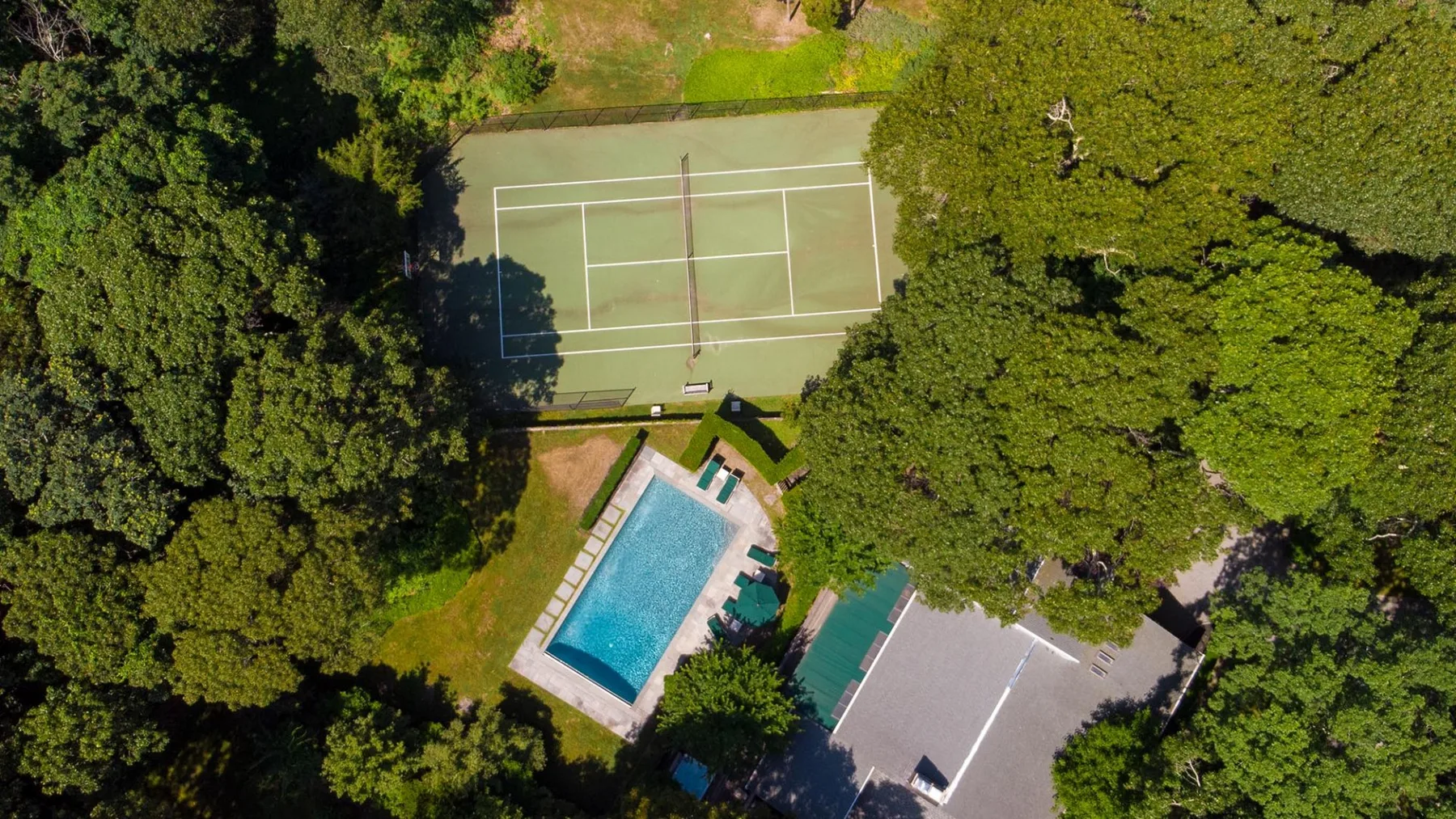 Una vista aérea muestra una piscina y una cancha de tenis entre árboles frondosos.