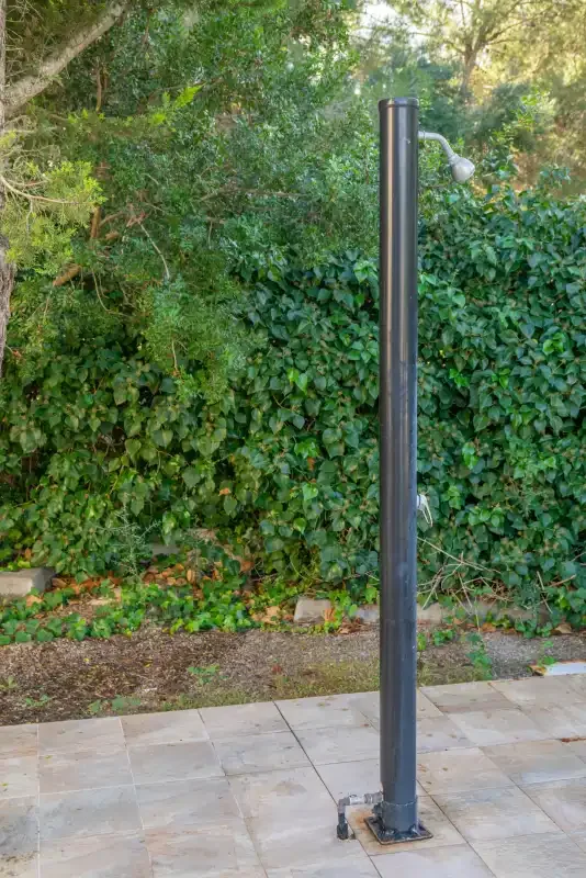 An outdoor patio with a freestanding shower beside a leafy hedge.