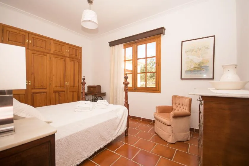 Un dormitorio con una cama, un armario de madera y un sillón junto a la ventana.