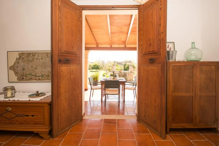 Unas puertas de madera abiertas dan paso desde una habitación con suelo de baldosas a un comedor con mesa y sillas.