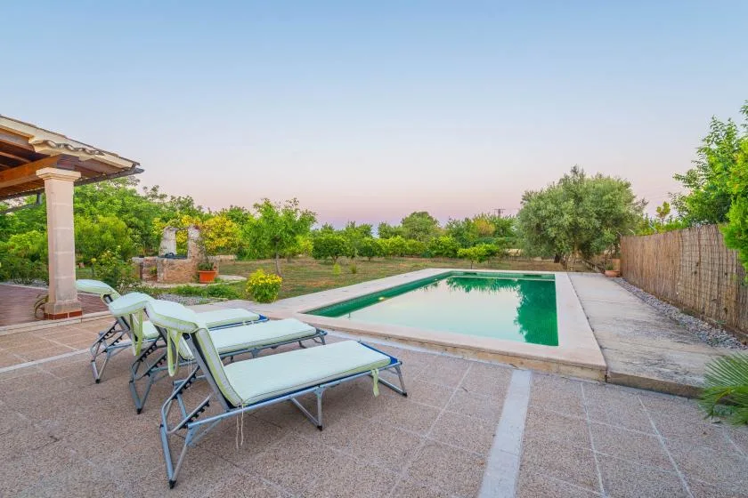 Una piscina junto a un patio pavimentado con asientos reclinables y árboles.