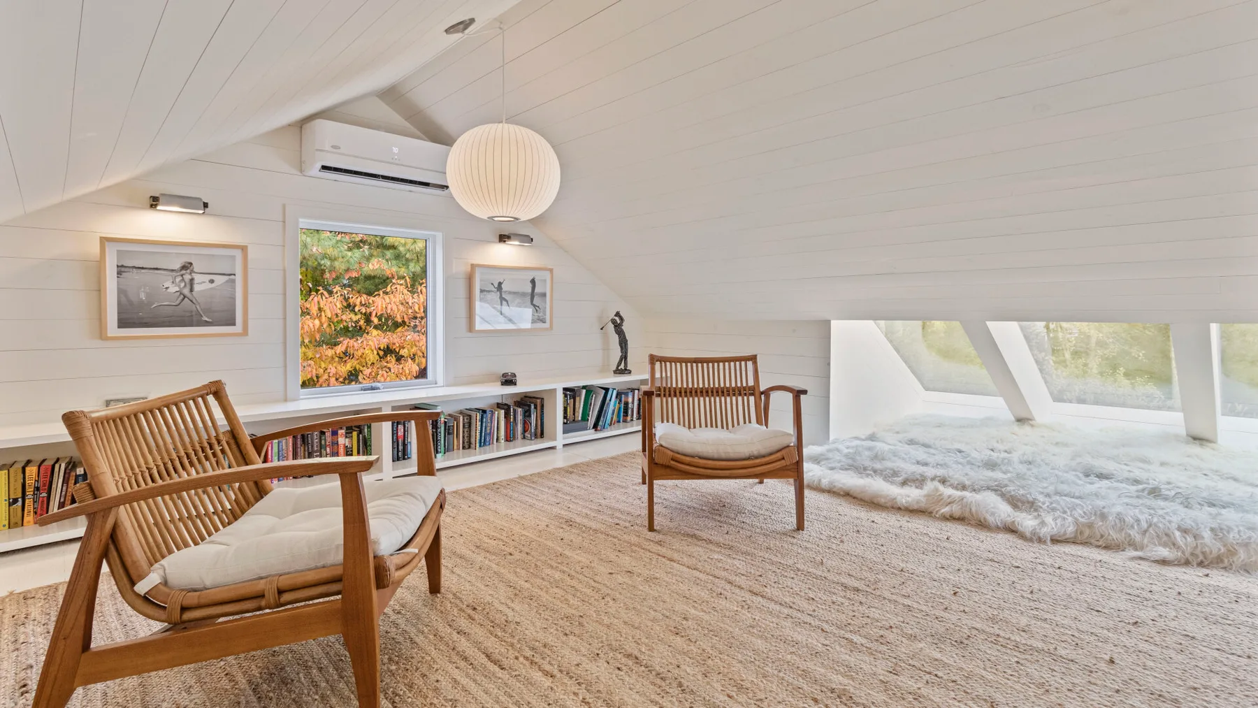 A bright attic sitting area has wooden seating, a low bookshelf, and sloped windows.