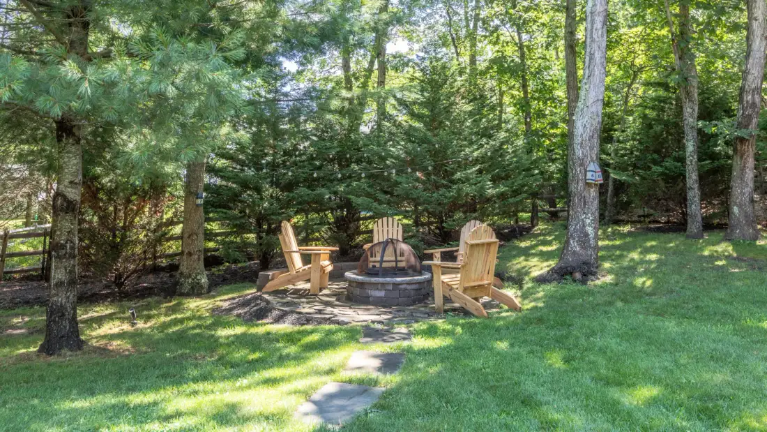 A grassy wooded outdoor area has seating arranged around a circular stone fire pit.