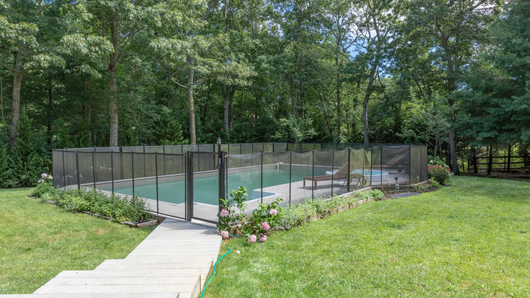 A fenced outdoor swimming pool sits in a grassy clearing among trees.