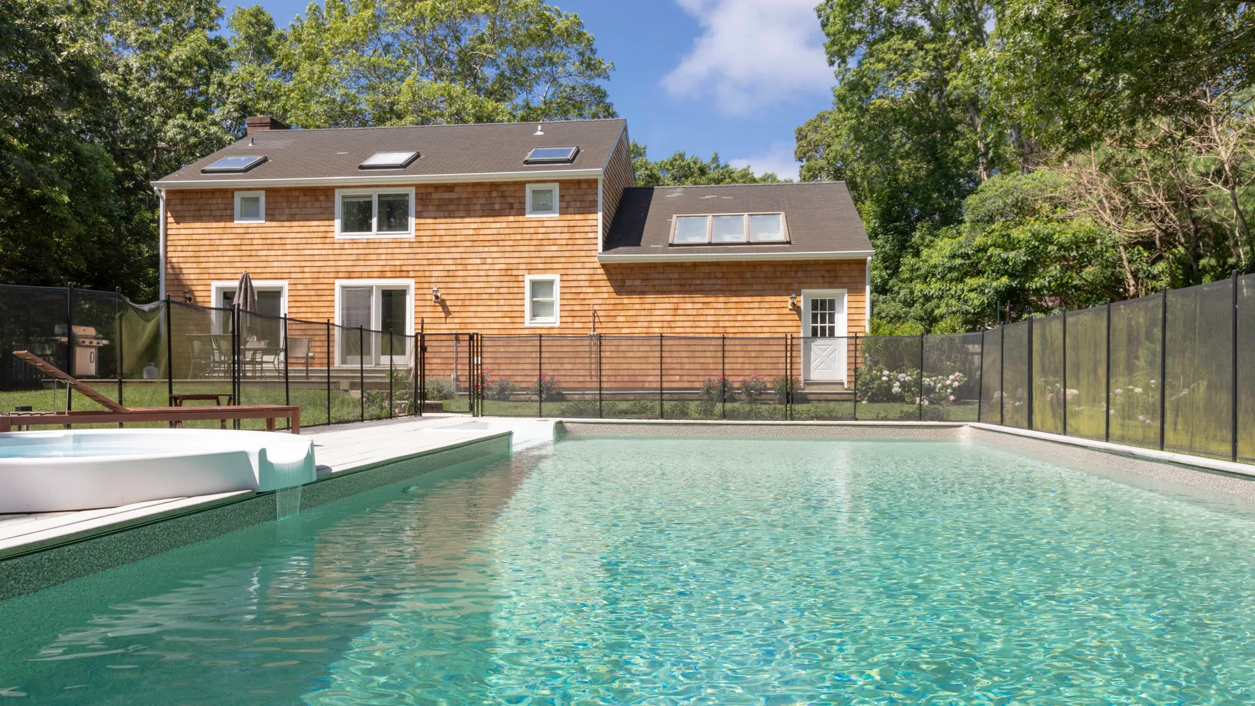 A swimming pool sits in front of a shingled house, enclosed by a fence.