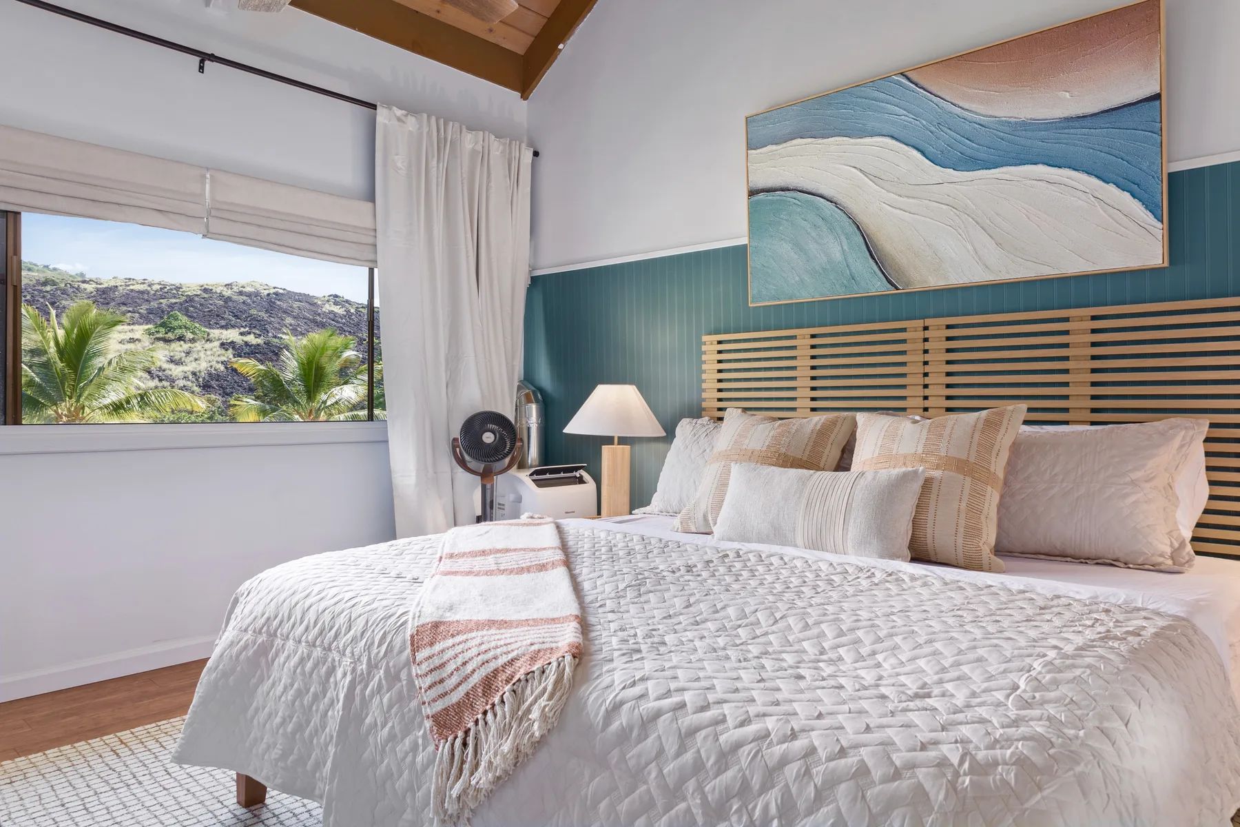 A bedroom with a neatly made bed, striped teal wall, and a window overlooking greenery.