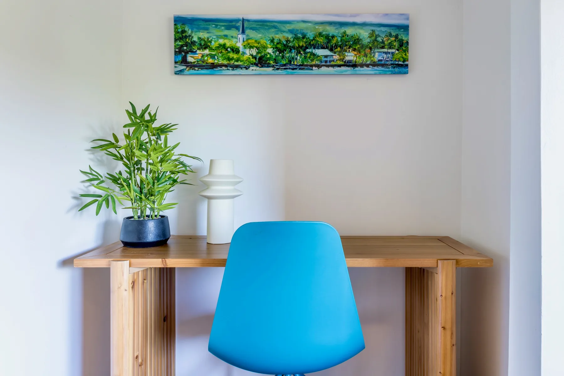 A desk and blue seating sit against a light wall with a plant and framed artwork.