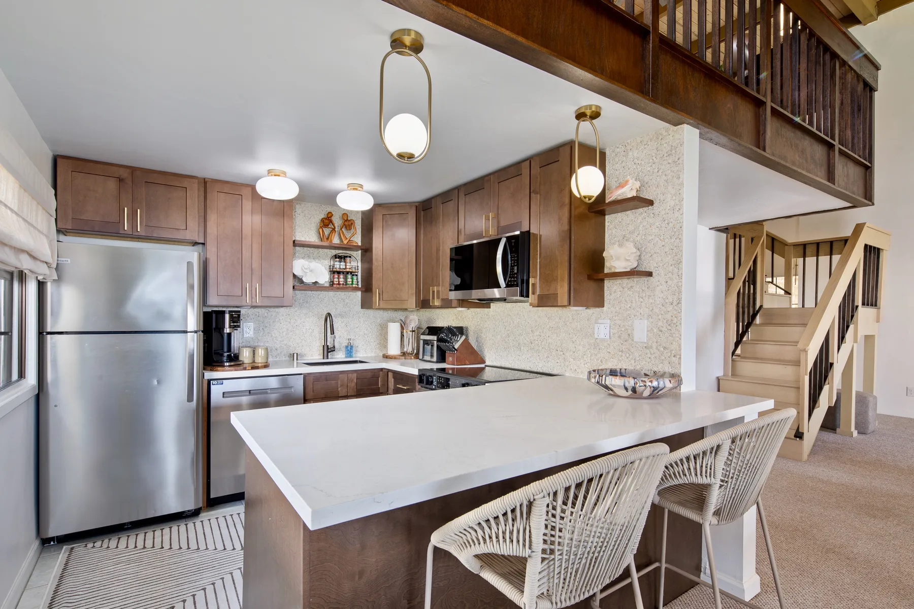 A kitchen with a large island and woven seating beneath a wooden loft railing.