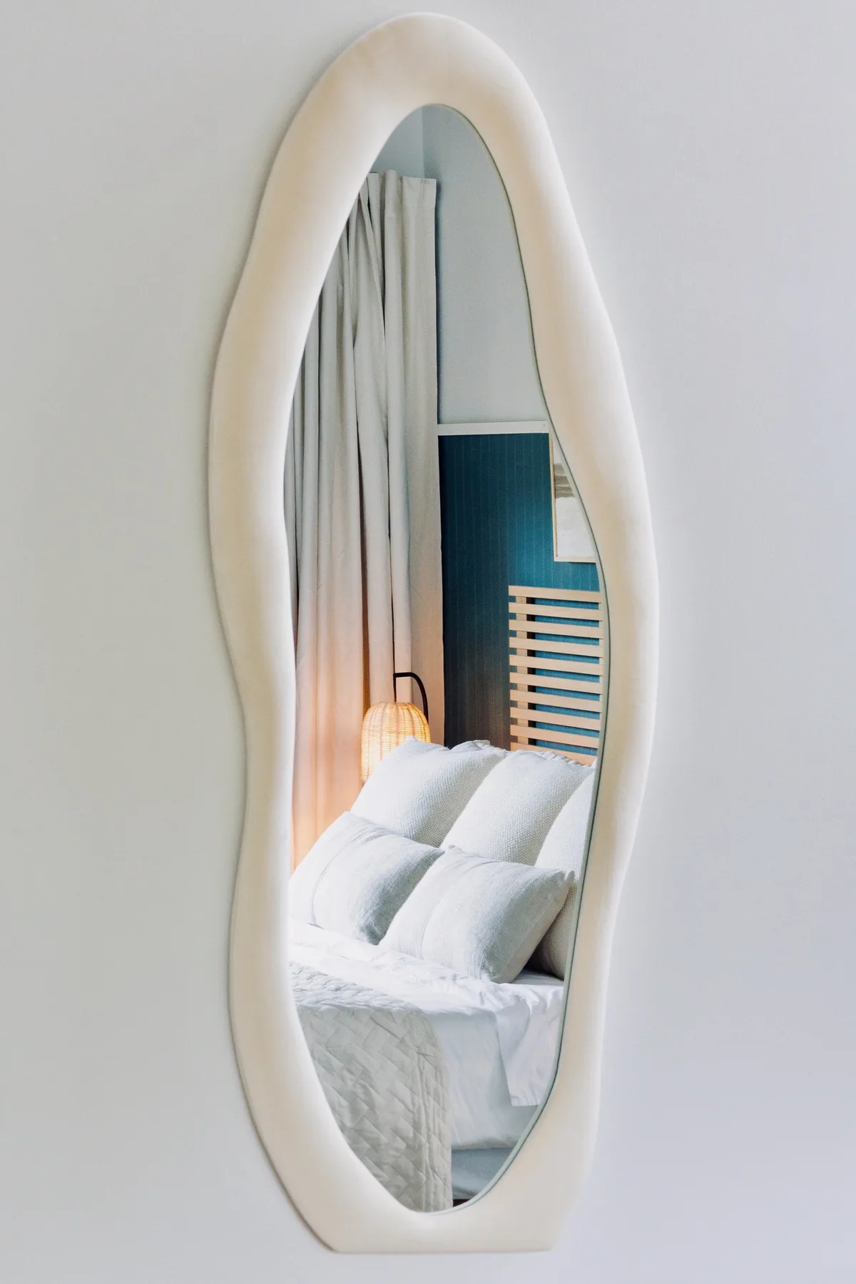A wavy wall mirror reflects a bed with white bedding and curtains.
