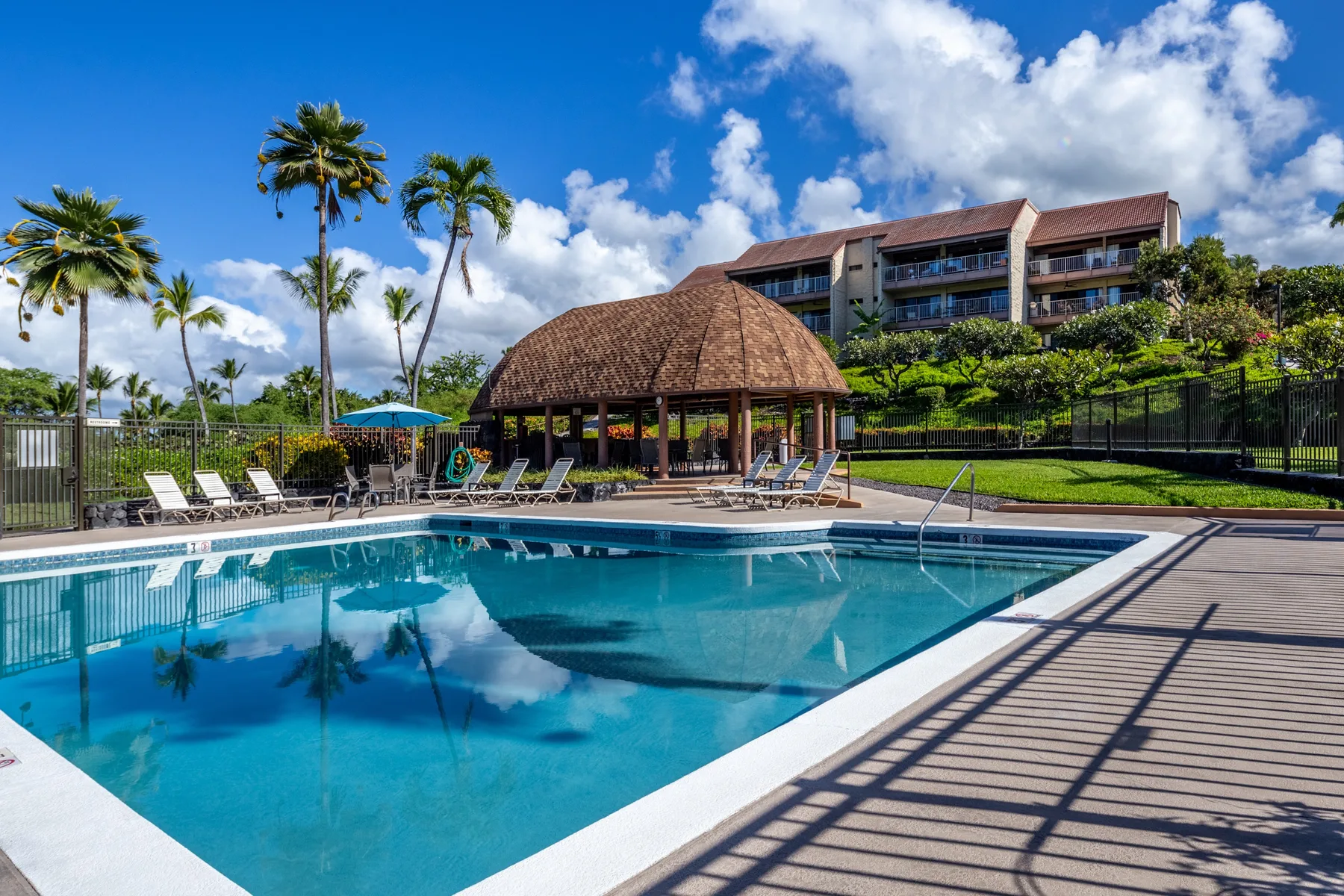 An outdoor swimming pool beside a covered pavilion and landscaped grounds.