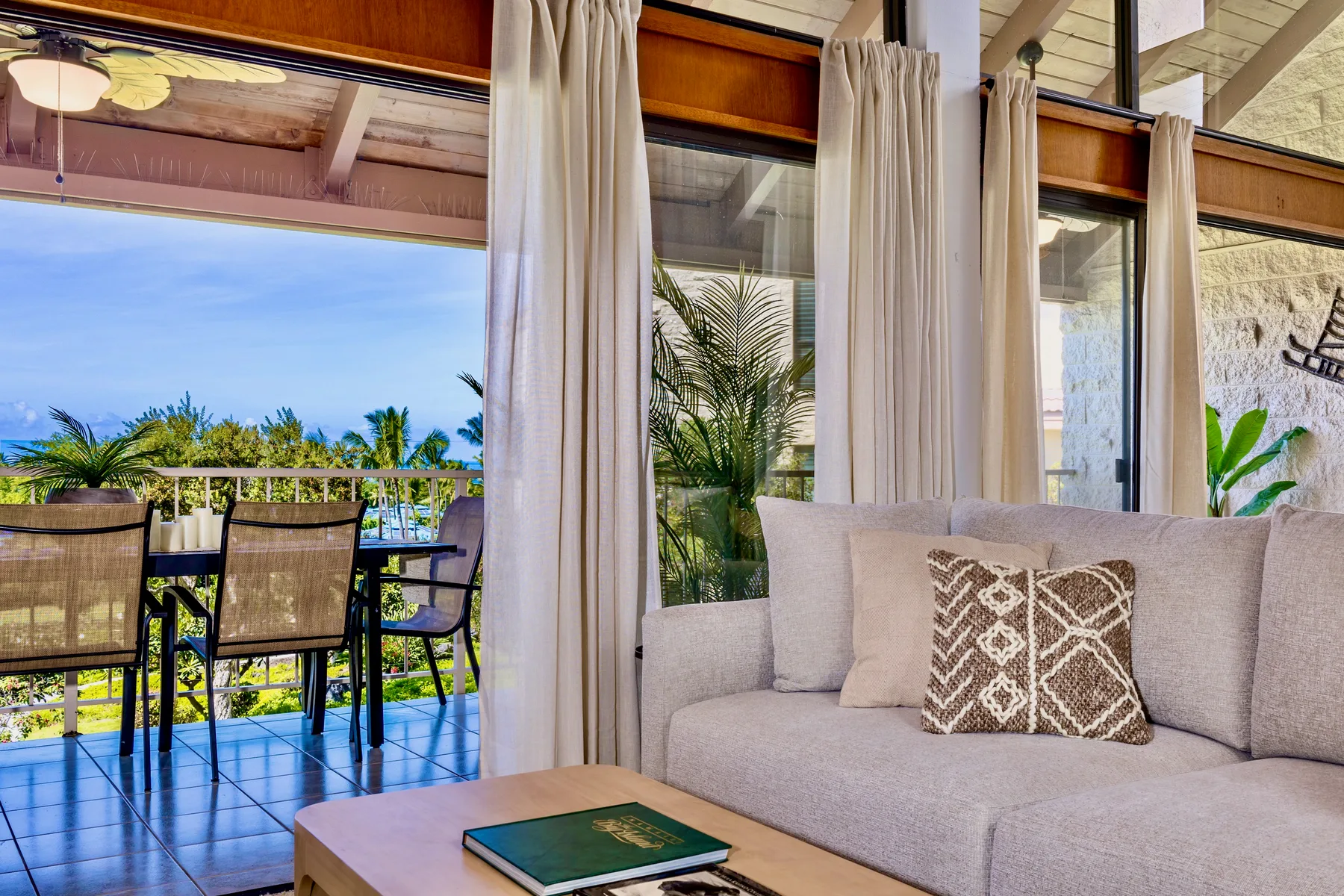 A bright living room opens onto a balcony with seating and tropical greenery.