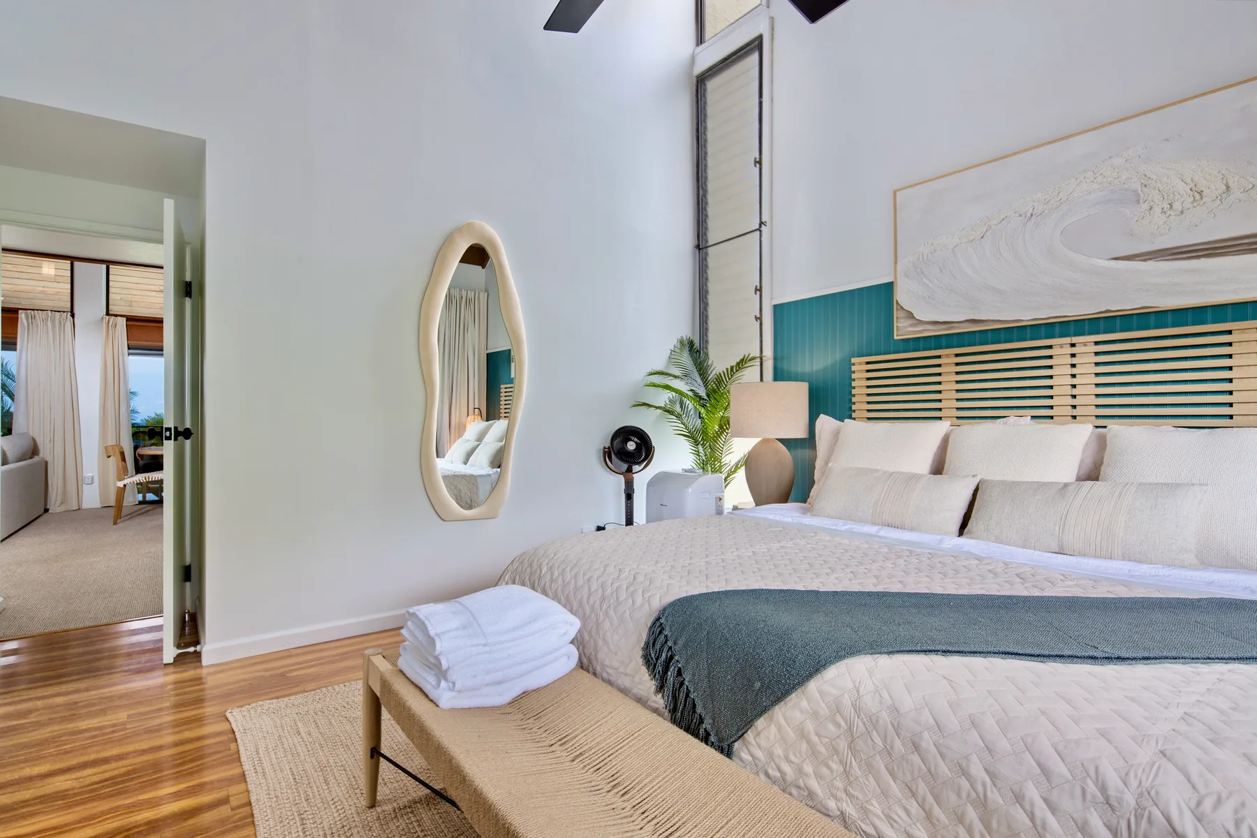A bright bedroom with a large bed, woven bench, and oval wall mirror.