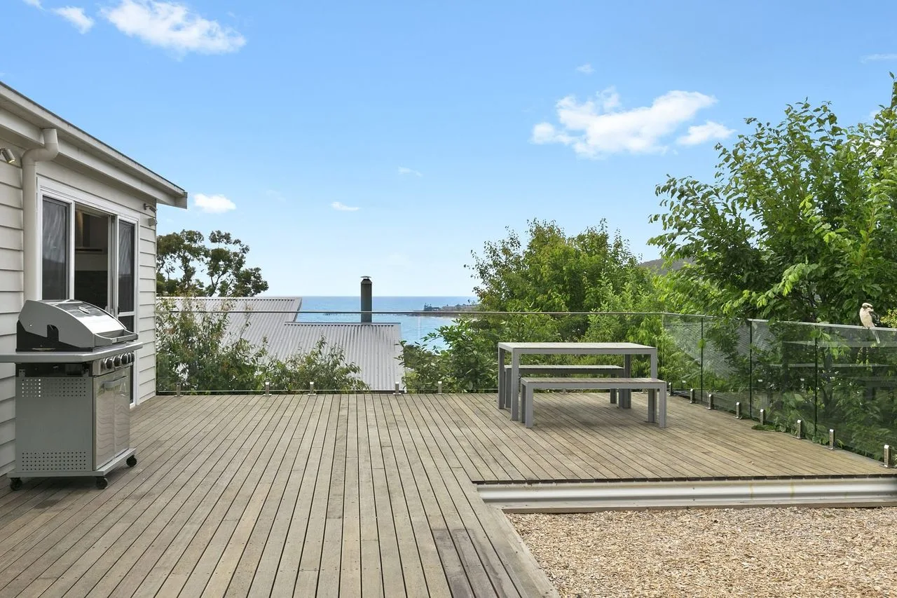 Una terraza exterior de madera da al agua, con asientos y una parrilla cerca.