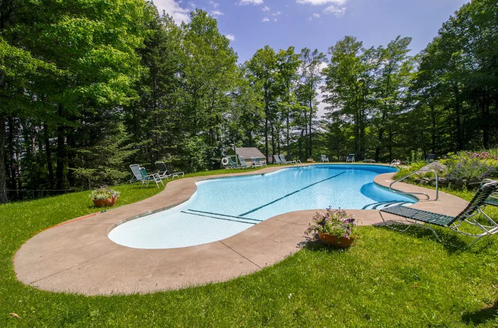 An outdoor swimming pool sits beside a grassy lawn and surrounding trees.