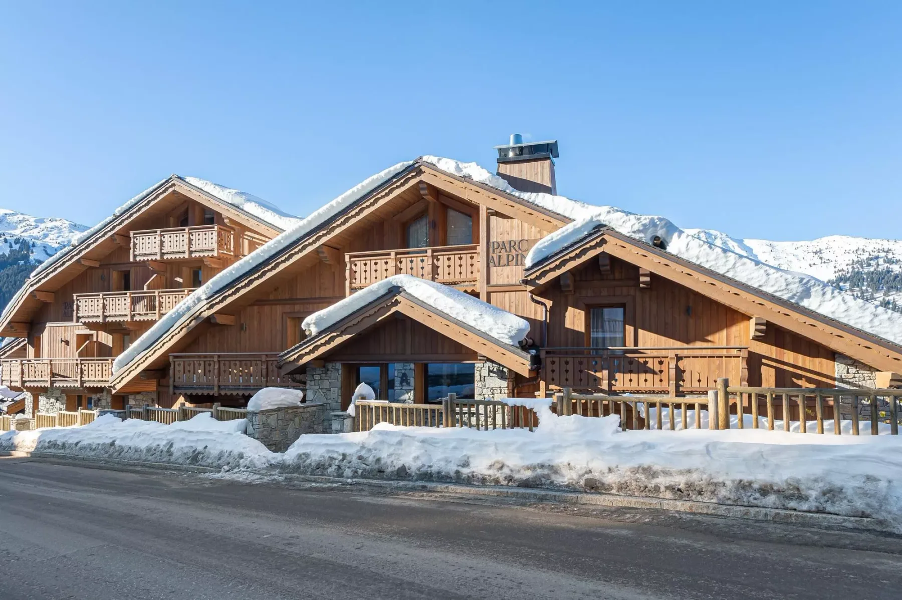 A large wooden lodge stands beside a snowy mountain road under a clear sky.