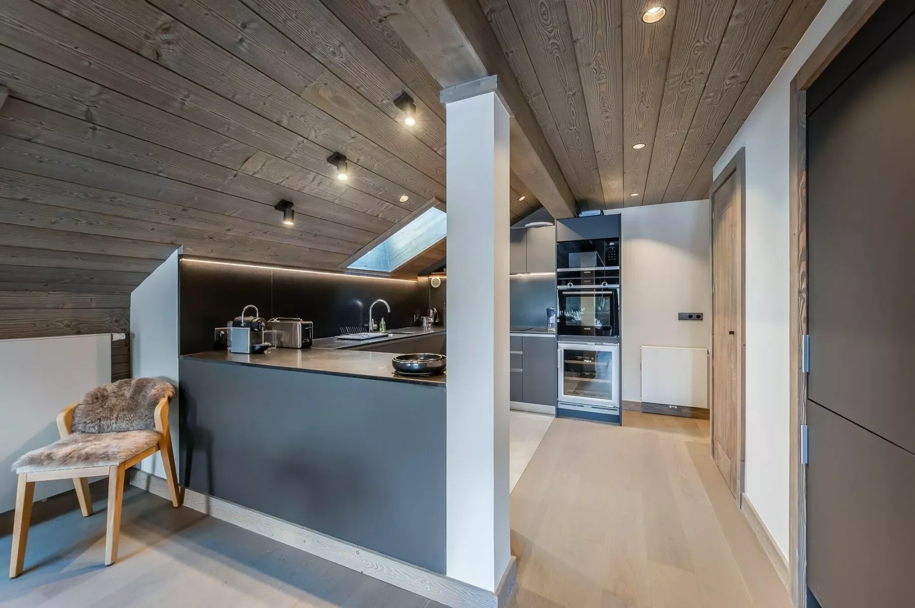 A modern kitchen with a dark island, built-in appliances, and a sloped wood ceiling.
