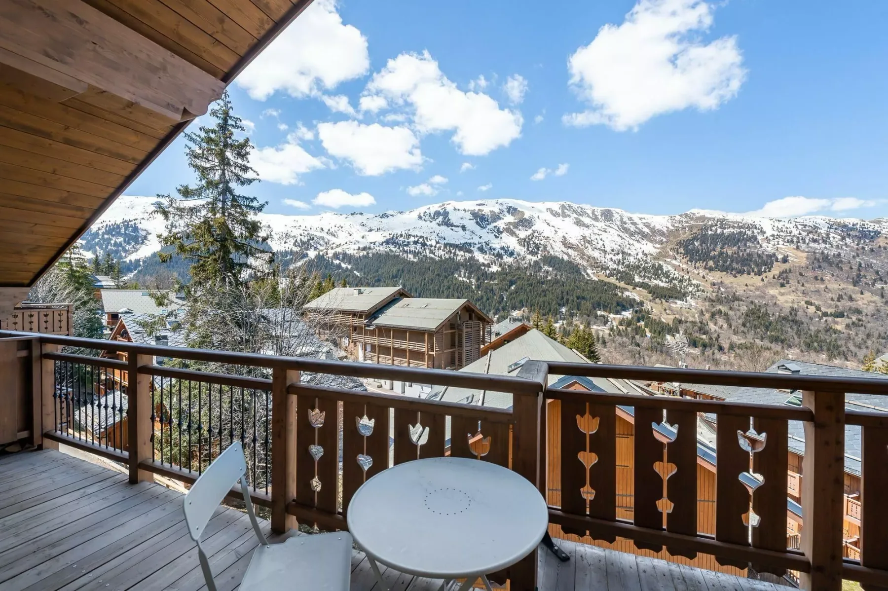 A wooden balcony overlooks mountain slopes with patches of snow beneath a blue sky.