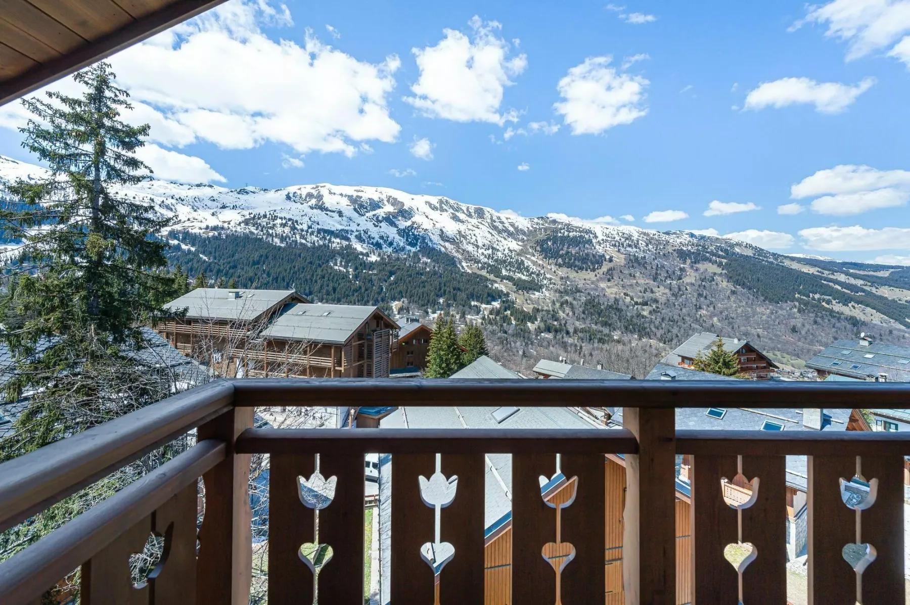 A wooden balcony overlooks snow-dusted mountains and buildings beneath a bright sky.