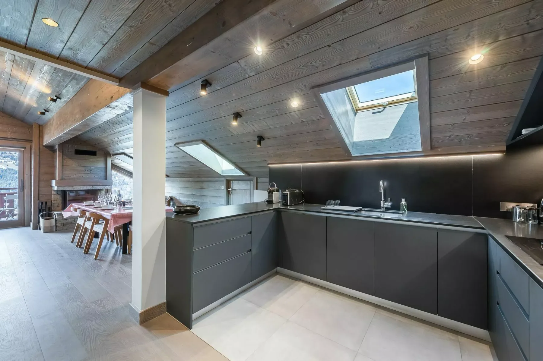 A modern kitchen sits beneath a sloped wooden ceiling with skylights.