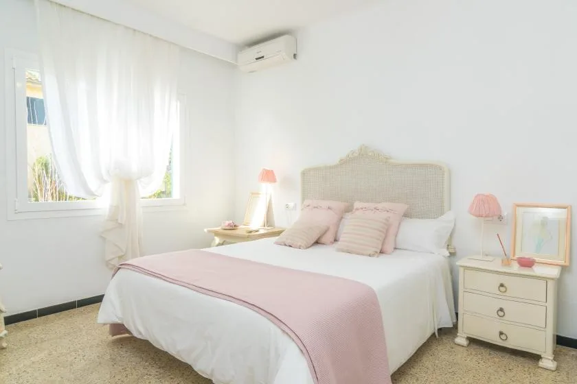 A bright bedroom with a white bed and soft pink bedding beside a curtained window.