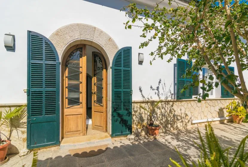 A white exterior wall has an arched wooden doorway with open teal shutters.