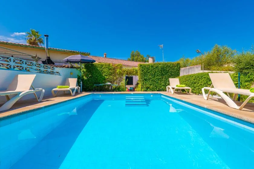A swimming pool with lounge chairs sits beside a hedge and low buildings.