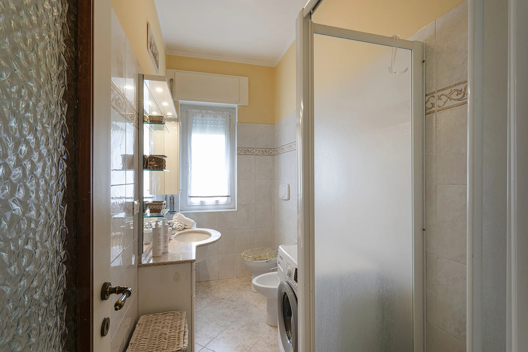 Un baño con azulejos, lavabo, ventana y ducha con mampara de vidrio.