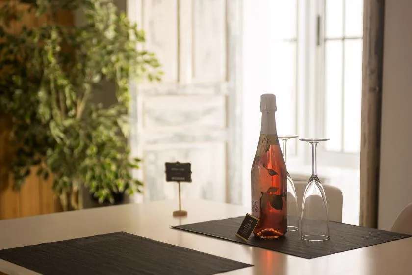 A dining table displays a bottle and glasses beside a bright window.