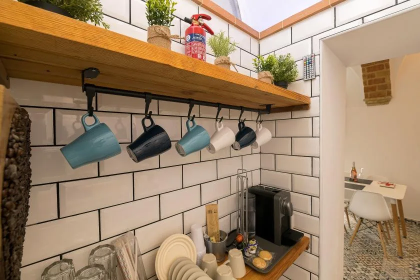 A tiled kitchen counter sits beneath a wooden shelf with hanging mugs.