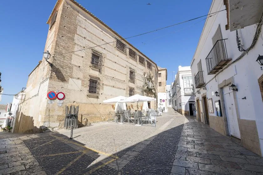 A sunlit cobbled street runs between pale buildings, with outdoor seating beneath umbrellas.