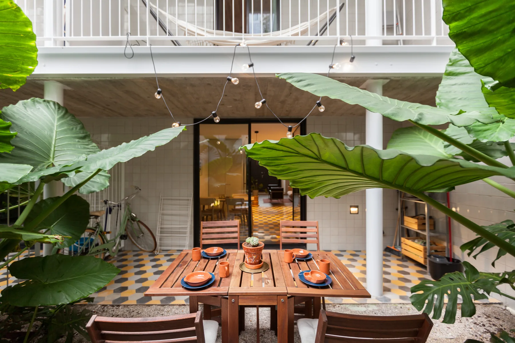 Un comedor en un patio con mesa de madera, suelo de baldosas de colores y plantas frondosas.